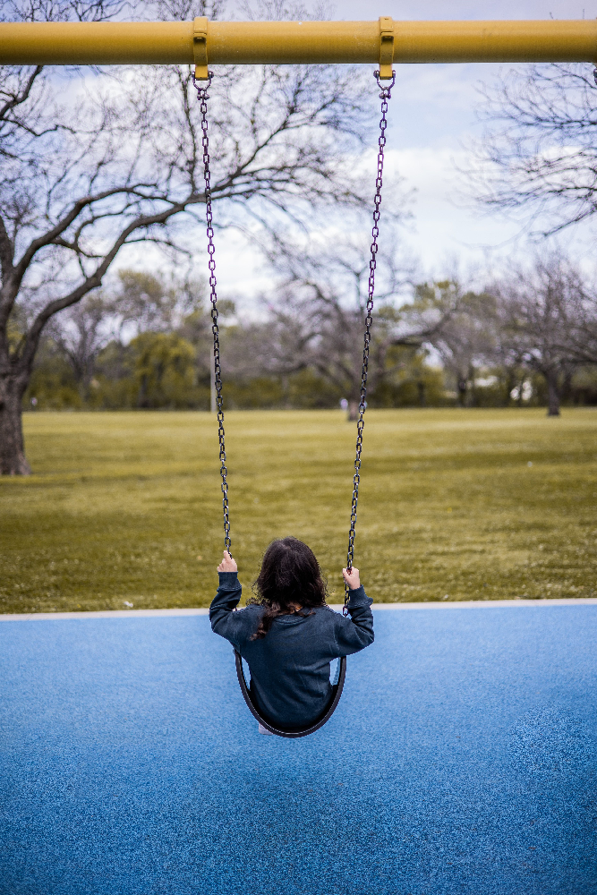 Child riding a traditional belt swing seat on a commercial playground swing set in a public park.